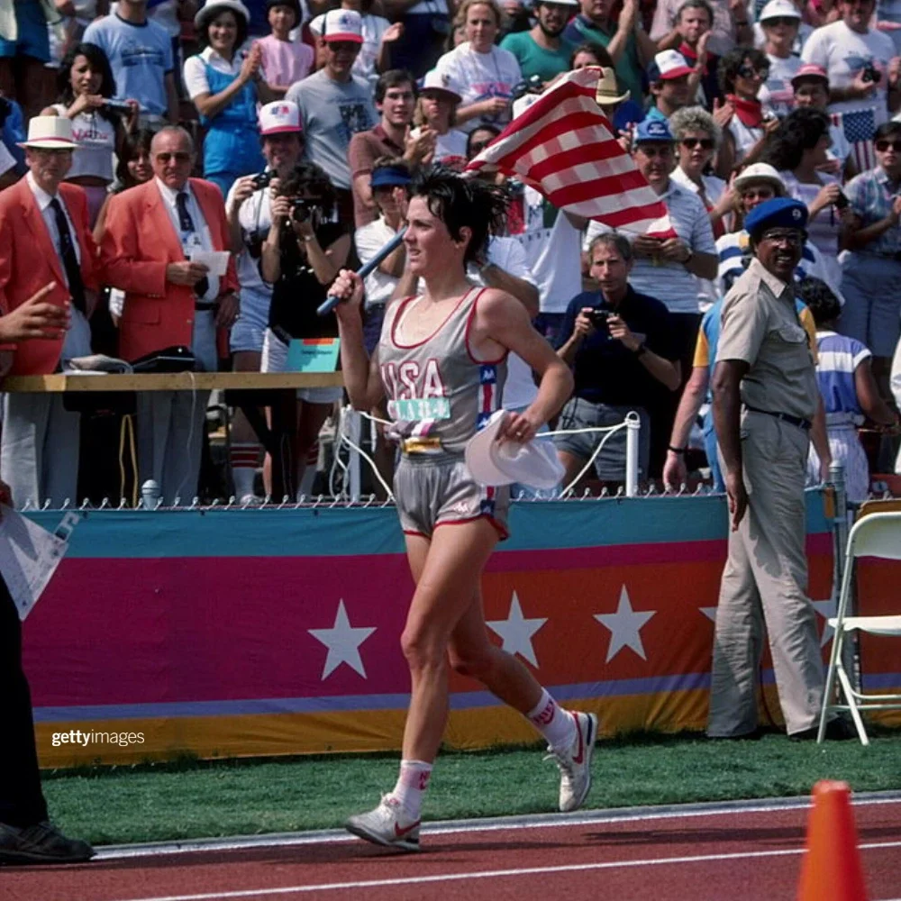 1984 Olympic Women's Marathon gold medalist Joan Benoit running in front of spectators holding the United States flag after winning the gold medal.