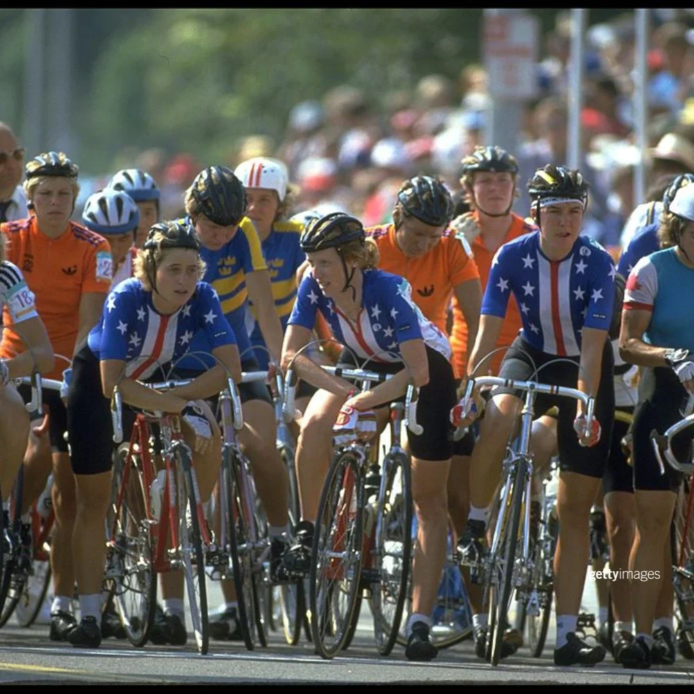 Members of the Team USA's women's cycling team in red, white and blue cycling outfits sitting on their bikes pre-race with cyclists behind them at the start of the road cycling race at the 1984 Games.