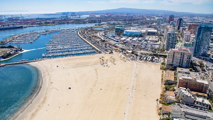 Fotografía aérea de Alamitos Beach en Long Beach. La arena está en el primer plano de la foto con edificios y condominios del centro de Long Beach en el lado derecho de la fotografía. El puerto deportivo costero de Long Beach, con barcos atracados, aparece sobre la sección de arena en la fotografía aérea. El Queen Mary, el Puerto de Long Beach y el Puerto de Los Ángeles aparecen en el fondo de la fotografía.
