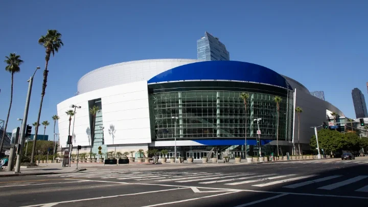 Fotografía del estadio del centro de Los Ángeles (Crypto.com Arena) en la que se ve la entrada principal con un diseño curvo y ventanas de cristal. A la izquierda de la fotografía, junto a la acera, se ven palmeras. 