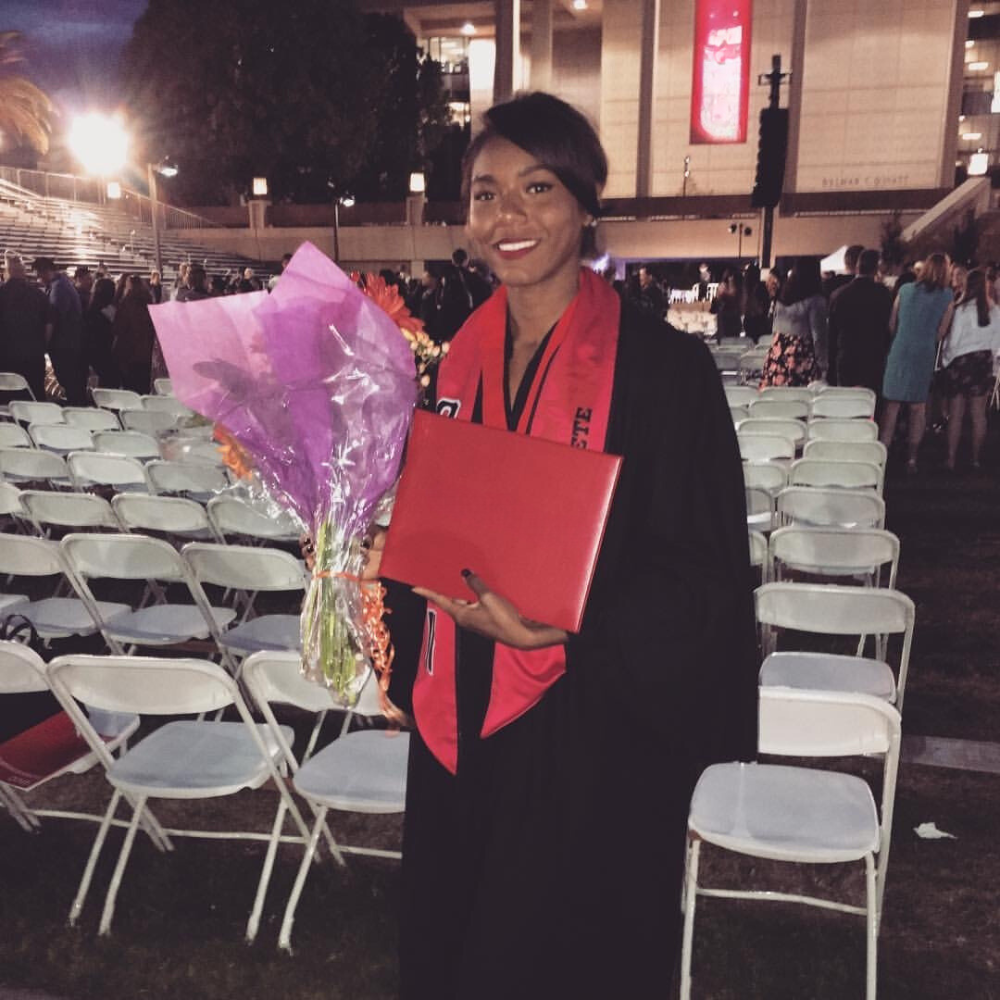 Sam Dirks standing in a black college graduation gown, smiling, holding flowers and diploma