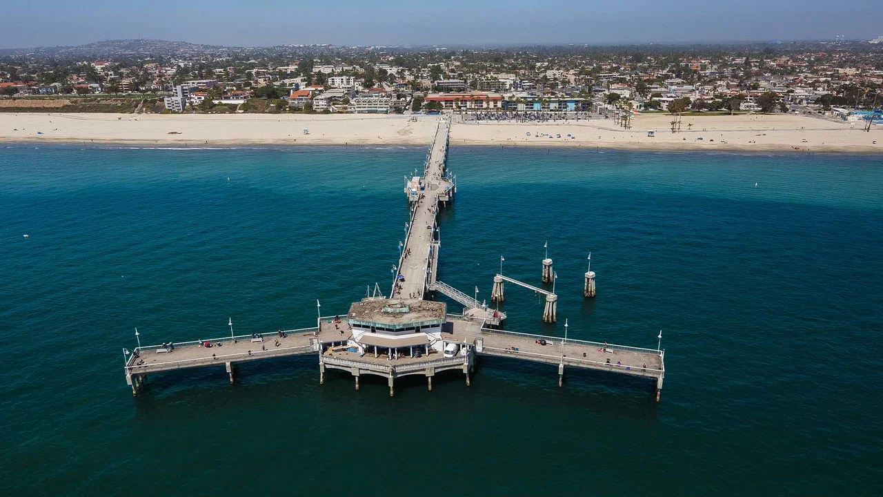 Fotografía aérea del Belmont Shore Pier en Long Beach, con el muelle extendiéndose en línea recta hacia el océano y luego desviándose hacia la derecha y la izquierda, con un pequeño edificio al final del muelle. Se puede ver a varias personas caminando sobre el muelle, y al fondo se observa una playa de arena, casas, edificios y un cielo azul.