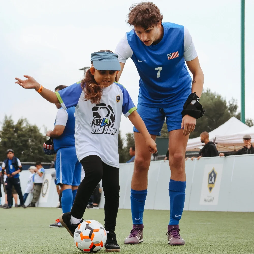 Una niña joven está pateando un balón de fútbol mientras un entrenador está detrás de ella apoyándola