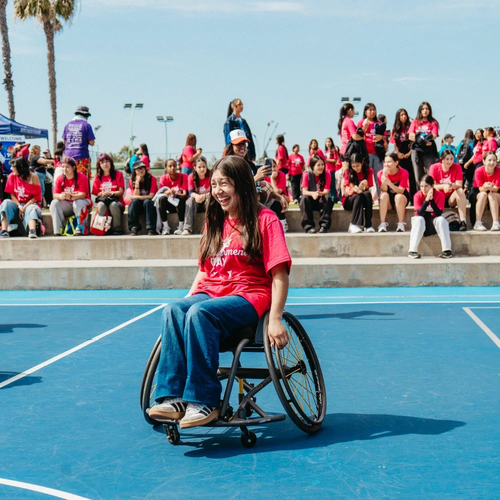 Una fotografía de una niña joven riendo y sonriendo mientras está sentada en una silla de ruedas en una cancha de tenis