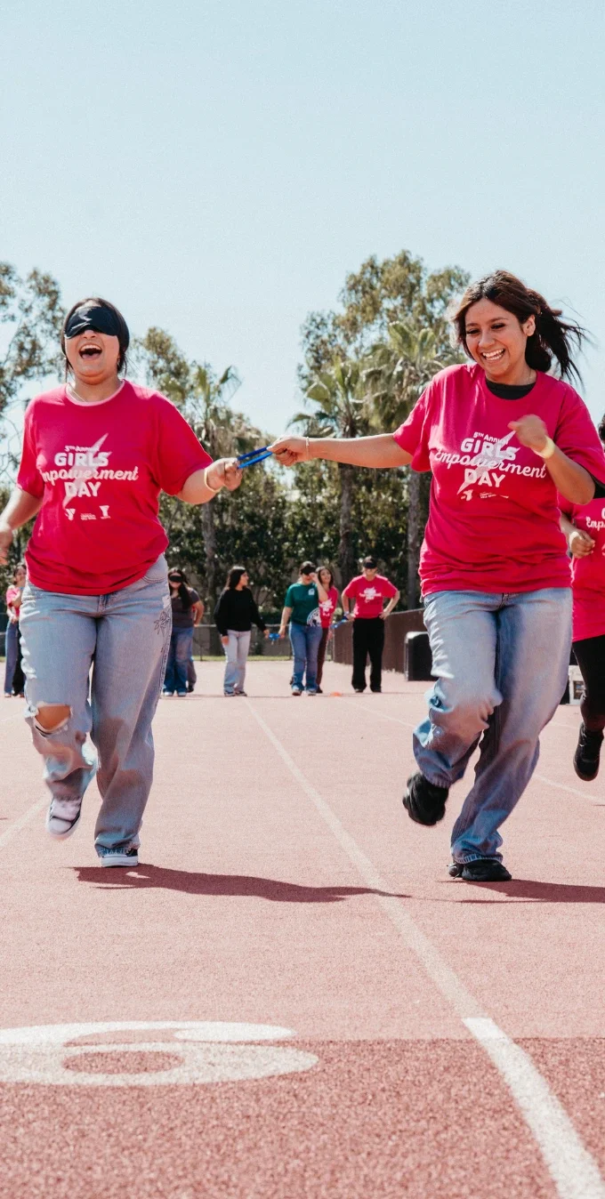 Fotografía de mujeres jóvenes corriendo en una pista al aire libre mientras una chica está con los ojos vendados e intenta pasar un relevo a otra chica que no está vendada