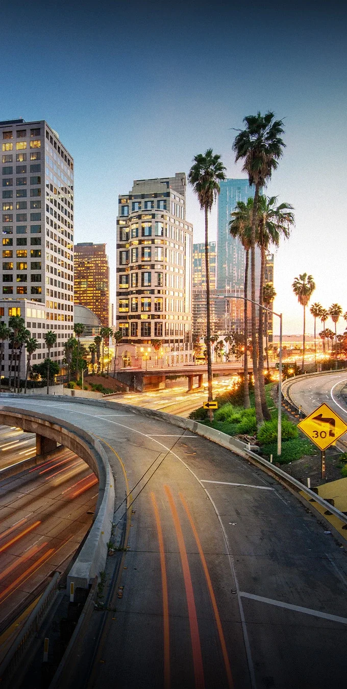Imagen de algunos rascacielos en el centro de Los Ángeles al atardecer, con una autopista borrosa en primer plano flanqueada por palmeras.