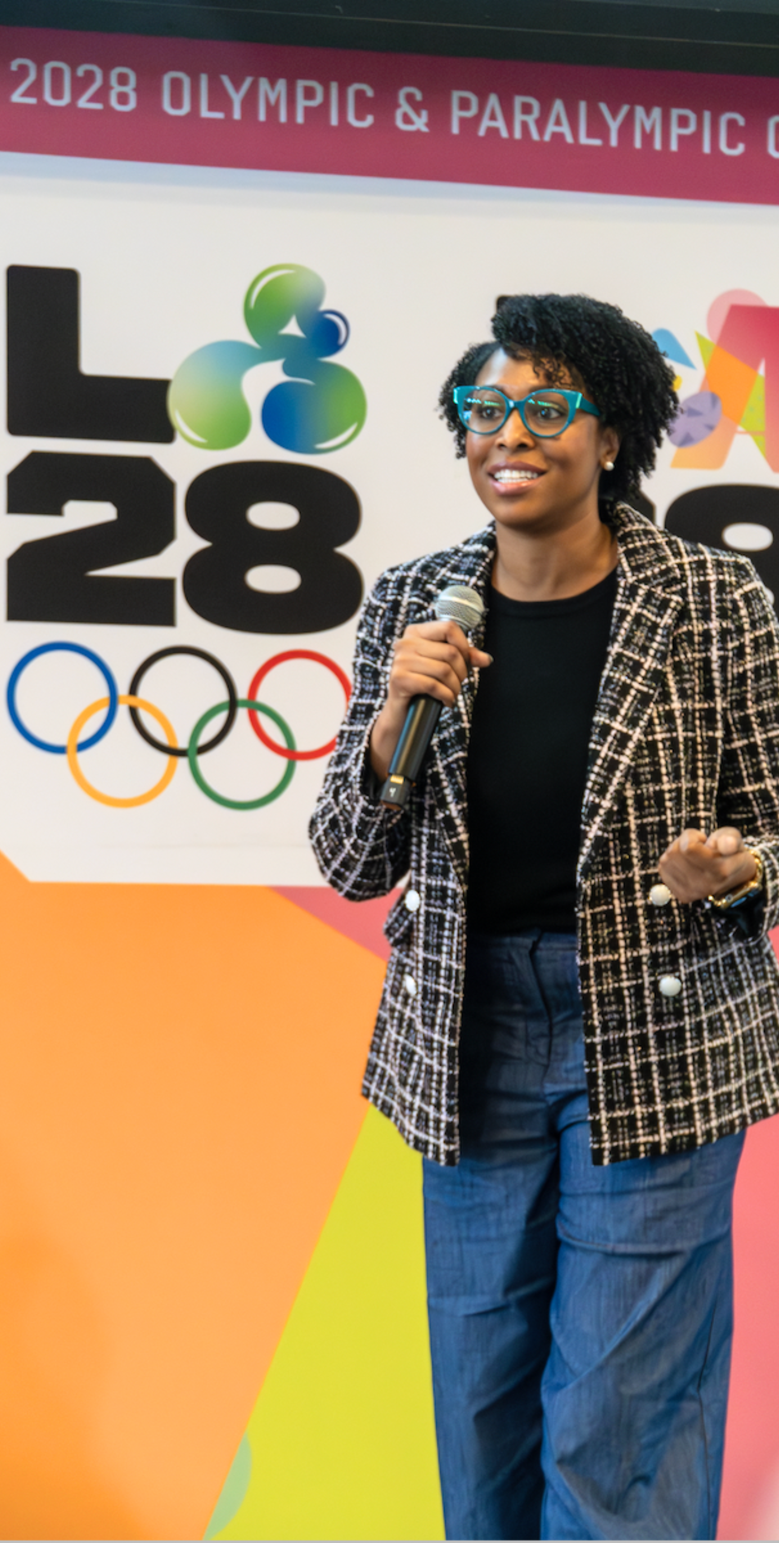 Esta fotografía muestra Porsia Curry, la Directora de Diversidad, Equidad, Inclusión y Pertenencia, sonriendo y hablando frente a un micrófono mientras ella está de pie frente a un fondo de LA28.