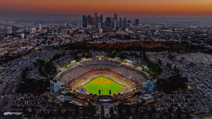 Una fotografía aérea del Dodger Stadium al atardecer. El campo del Dodger Stadium está iluminado mientras los aficionados están sentados. El aparcamiento que rodea el estadio está lleno de coches y al fondo se ve el perfil del centro de Los Ángeles.