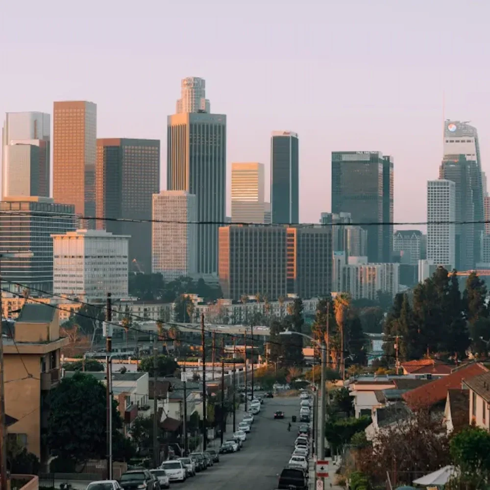 El horizonte del centro de Los Ángeles visto al atardecer desde una calle en una colina bordeada de autos y edificios de apartamentos. También se observan árboles y postes de electricidad en la imagen. 