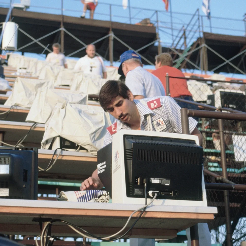 Fotografía de un hombre reparando una computadora del sistema de mensajería electrónica durante los Juegos de 1984; al fondo, en una sede al aire libre, se ven filas de computadoras y otras personas trabajando en ellas.