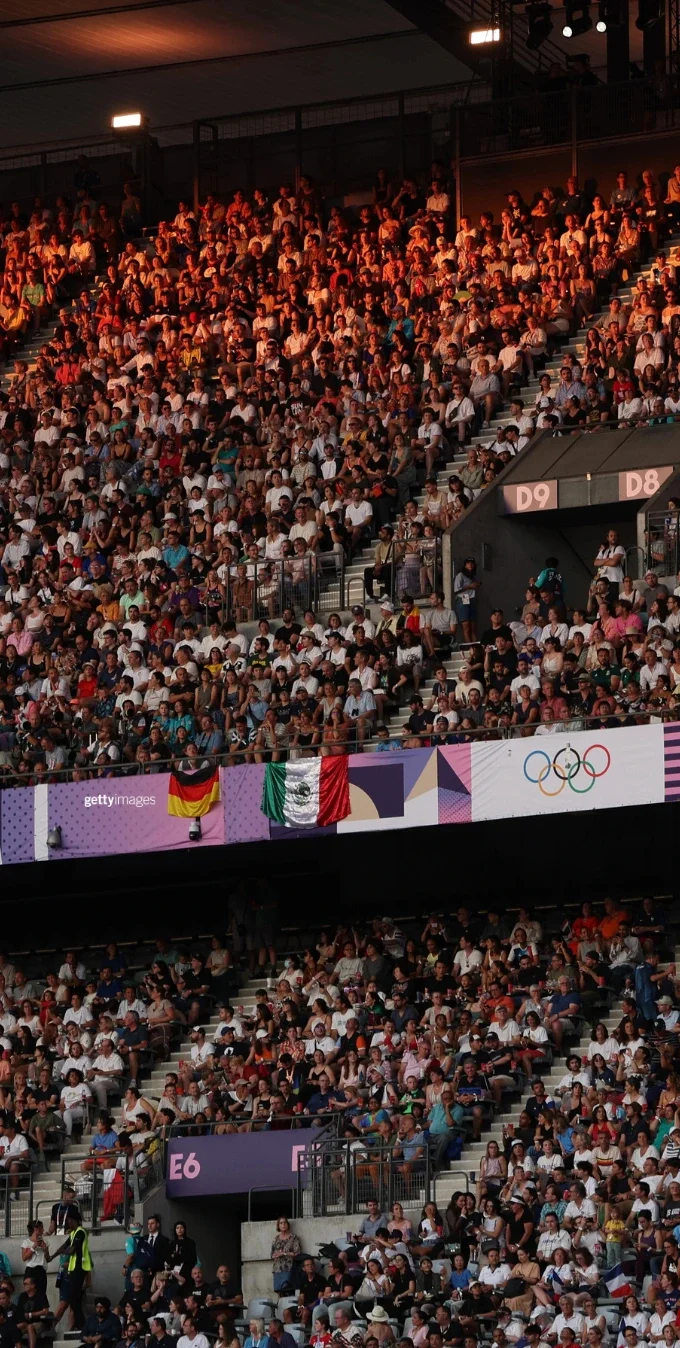 Aficionados en las gradas de un estadio techado durante los Juegos Olímpicos de París 2024. Las banderas de Alemania y México cuelgan de una de las barandillas junto a señalización con los aros olímpicos.
