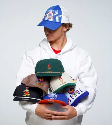 Hombre rubio con una gorra de la Colección Herencia Olímpica, vestido con una sudadera con capucha y sosteniendo varias gorras de la Colección Herencia Olímpica