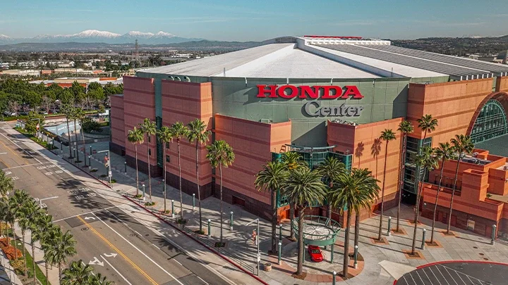 Una fotografía de Honda Center desde un ángulo posterior. Se ve la fachada bronceada del edificio con paneles verde oscuro. Las palmeras y una calle bordean el edificio por el lado izquierdo. Un coche rojo está estacionado bajo un gazebo verde oscuro frente a la entrada del estadio. Un grupo de palmeras se muestran en frente de la entrada de la arena. 