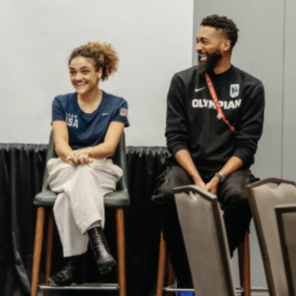 A photograph of Olympic gymnast Laurie Hernandez wearing a navy blue Team USA shirt sitting next to Jason Richardson who was wearing a black sweater with the word Olympian on it,  on stools during a panel talk