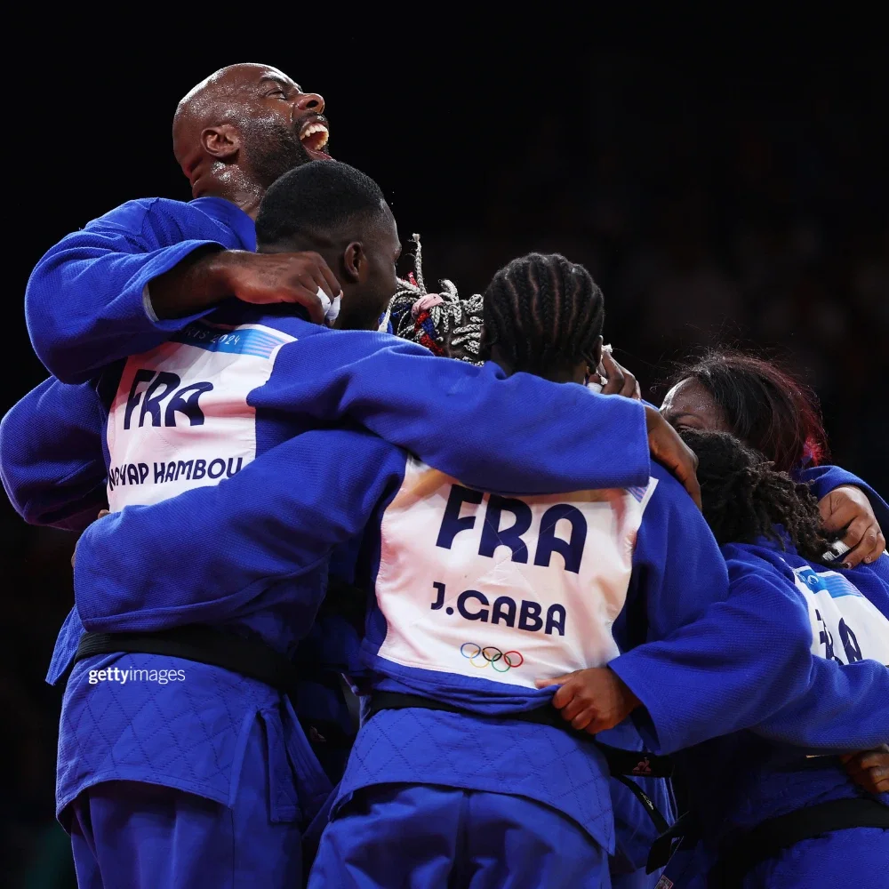 Un grupo de atletas olímpicos de judo celebrando y abrazándose en círculo; el equipo está formado por hombres y mujeres.”