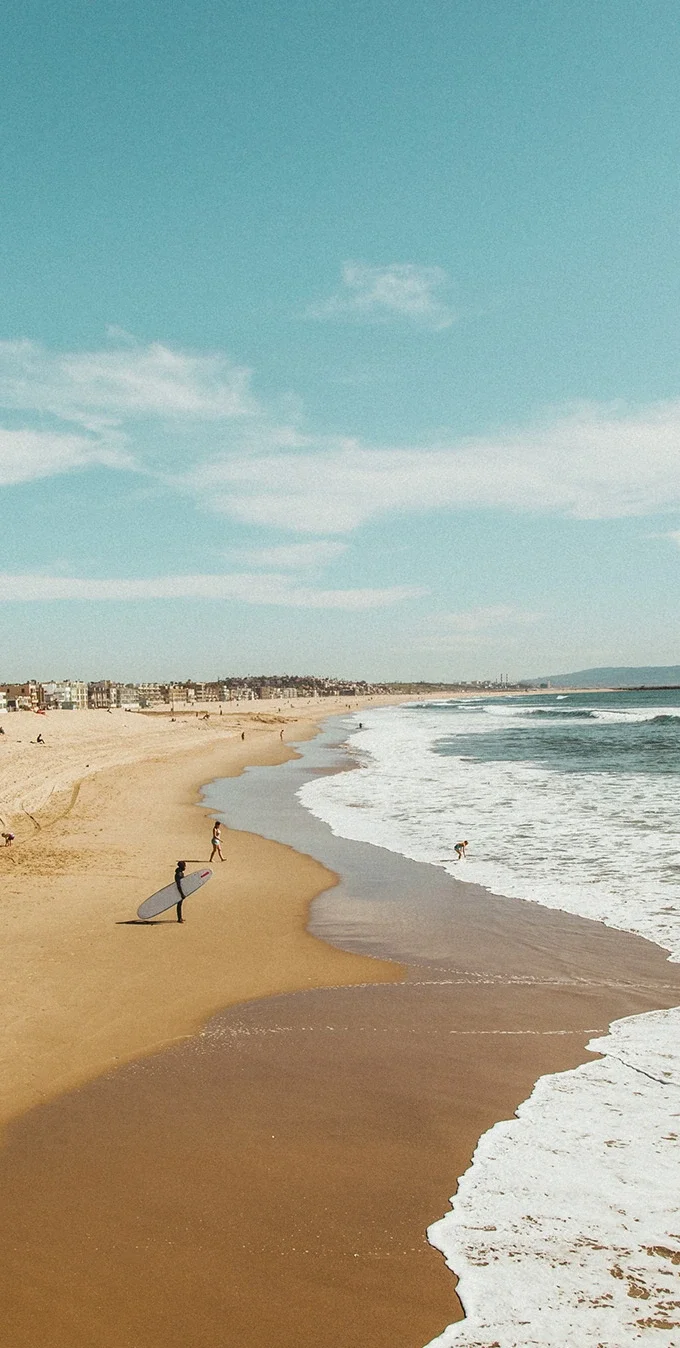 Fotografía de una playa con arena marrón y olas de color azul claro con espuma batiente en la orilla. Hay un surfista sosteniendo una tabla de surf de pie en la arena y una mujer observando a un niño jugar en el agua. Al fondo, casas bordean la orilla de arena y hay personas sentadas en la arena.