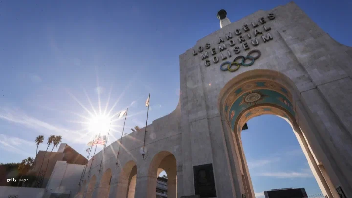 LA Memorial Coliseum se muestra desde el frente durante el día. Banderas, incluyendo la bandera estadounidense, ondean al lado izquierdo de la fotografía, con palmeras flanqueando el Coliseum en ese mismo lado. También hay una marca de agua de Getty Images en la parte inferior izquierda de la foto.