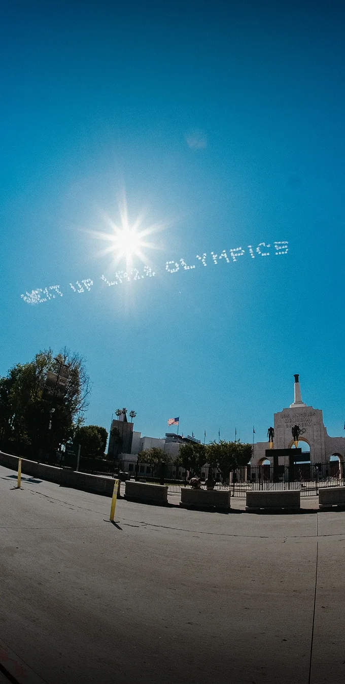 Imagen del LA Memorial Coliseum con un cielo azul de fondo y las palabras "Next Up LA28 Olympics" escritas en el cielo por un avión.