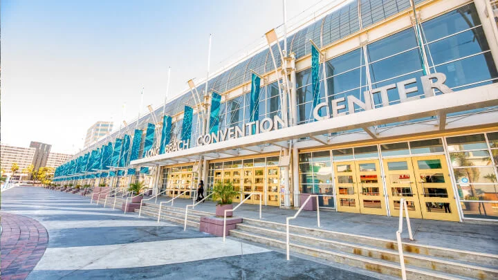 Fotografía del Convention Center en Long Beach, un edificio largo y rectangular con puertas amarillas y una fachada de cristal arqueada con suelo de hormigón en el exterior, escalones y barandillas blancas y banderas azules colgando de la fachada. Sobre la entrada se lee "Long Beach Convention Center" en grandes letras plateadas.