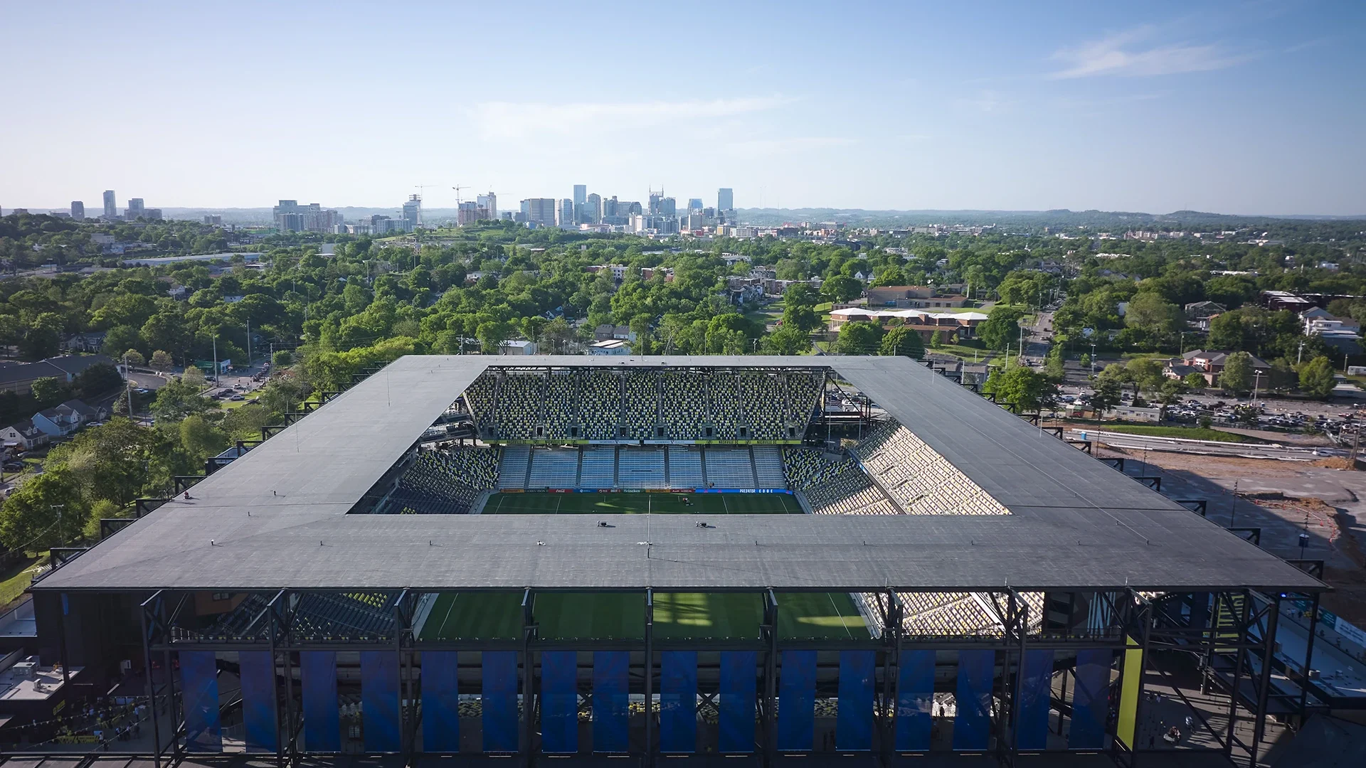Vista aérea de Nashville Stadium, con un techo al aire libre y las gradas para los aficionados en el centro del estadio. Al fondo se observan árboles, casas y los edificios del centro de Nashville.