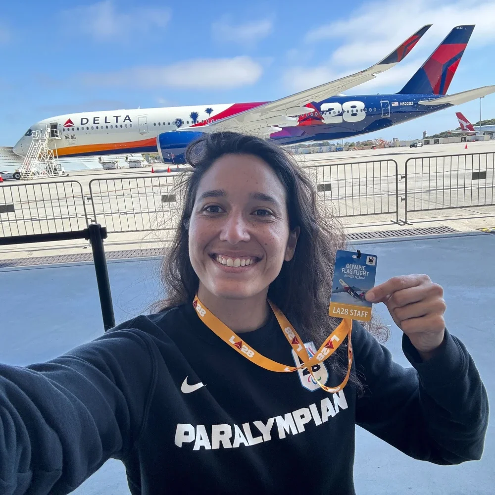 A photograph of Natalia Mayara in a black sweater that says Paralympian while holding up a lanyard badge in front of the Delta plane for the Delta Olympic flag flight