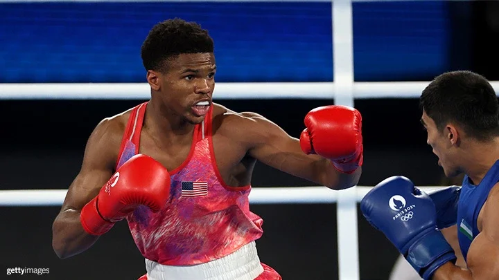 A photo of a Team USA boxer wearing a red, white and blue tie-dyed boxing shorts and a tank top with the American flag on the left chest throwing a left punch at his opponent
