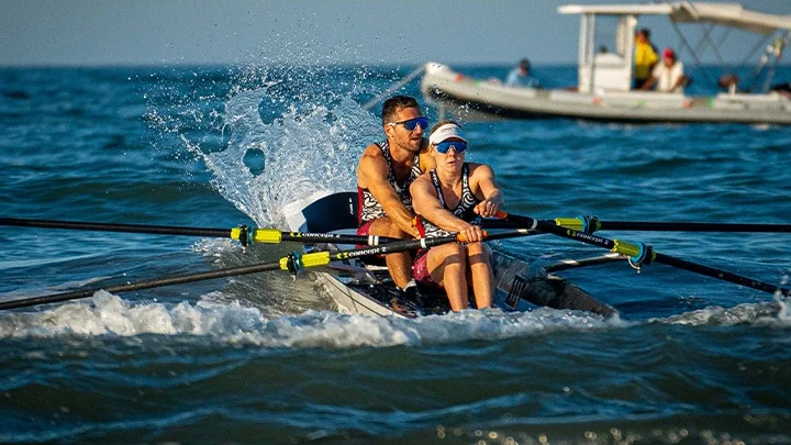 Fotografía de un equipo mixto formado por una remera delante y un remero detrás en el océano, con una barca blanca con varias personas a bordo al fondo.