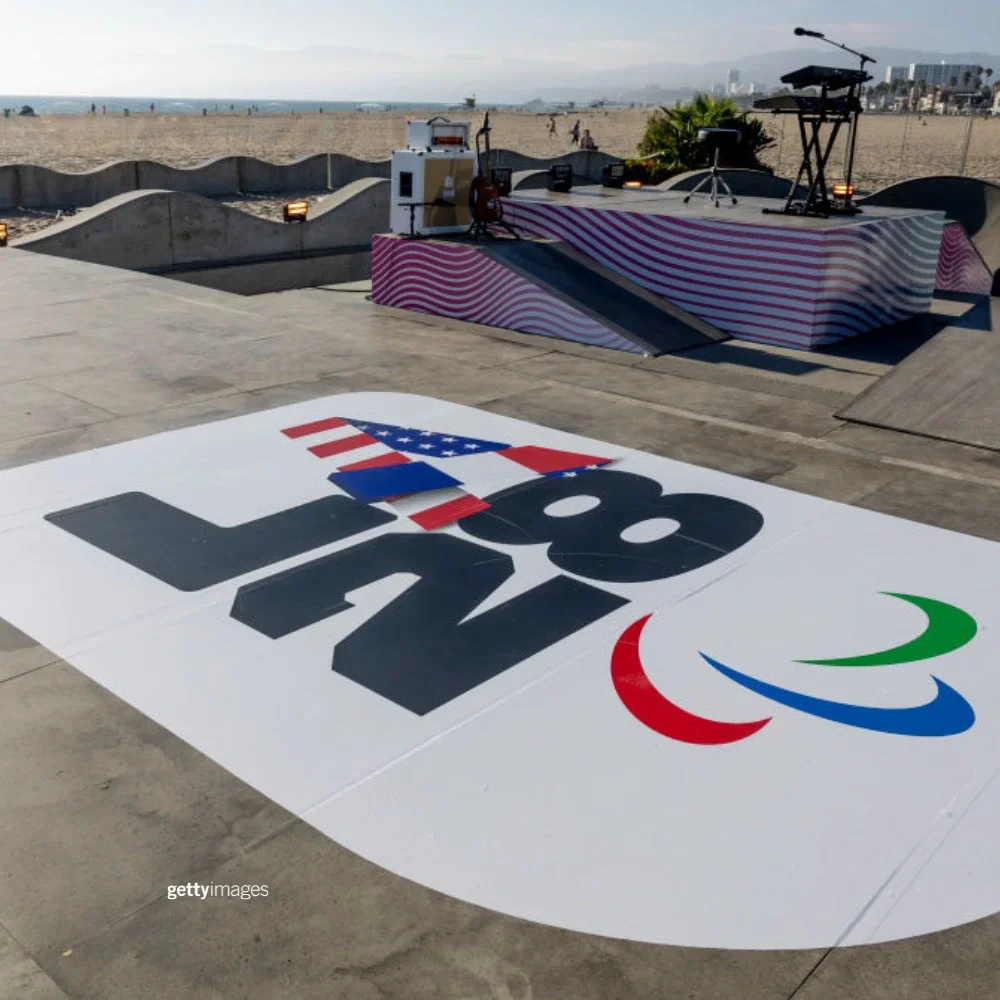 Foto del emblema de la entrega de los Juegos Paralímpicos LA28 en un gráfico en el suelo del parque de patinaje de Venice Beach, con los agitos paralímpicos debajo. Al fondo se ven un soporte de teclado vacío y micrófonos