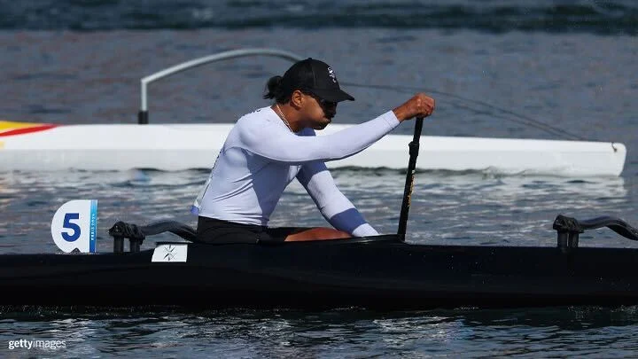 Una toma dinámica de una carrera de canoas en un cuerpo de agua tranquilo con varias atletas femeninas remando enérgicamente. Las canoas están agrupadas con marcadores de carril visibles numerados del 1 al 7 de derecha a izquierda. La atleta en primer plano, vestida con una camiseta blanca de manga larga y gafas de sol, está en el kayak número 7. Los atletas están en pleno remo, generando salpicaduras al remar vigorosamente en el agua.