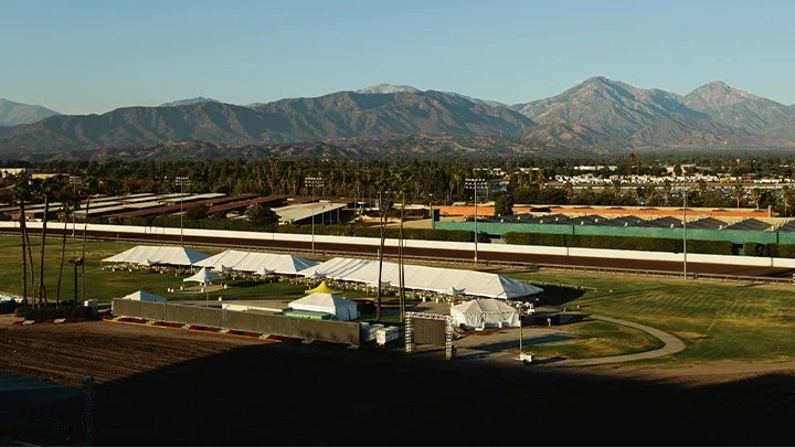 Fotografía del recinto Fairgrounds en Pomona con carpas blancas en el centro de la fotografía sobre el césped. Las luces del estadio están detrás de las carpas blancas y las montañas de San Gabriel están en el fondo.