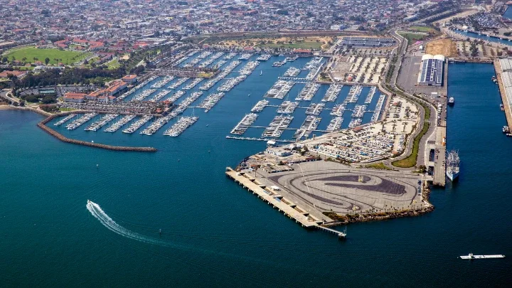 Una fotografía aérea del Puerto de Los Ángeles que muestra la costa azul del océano y una marina llena de barcos, con una plataforma de aterrizaje para helicópteros en primer plano, y al fondo, un paisaje urbano con casas y edificios.