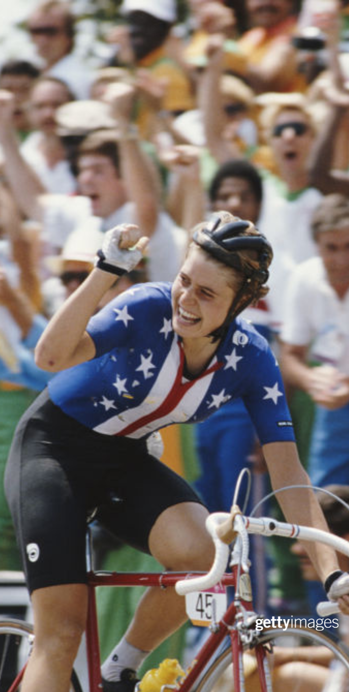 Rebecca Twigg in a Team USA Cycling uniform raising her right hand into a fist while riding a bike in the women's cycling event at the 1984 Games.