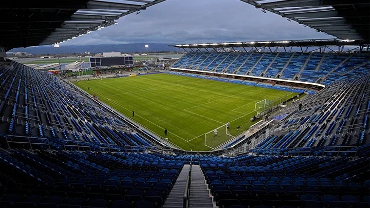 Vista interior de un estadio con filas de asientos azules vacíos y una vista inclinada de la cancha de fútbol verde, donde varias personas se encuentran de pie a lo largo de los bordes.
