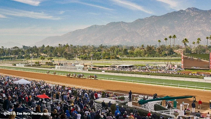 Fotografía de Santa Anita Park con jinetes montados en caballos que corren en la pista de tierra mientras los espectadores están sentados en las gradas en el primer plano de la fotografía. Las montañas de San Gabriel y las palmeras aparecen en el fondo de la fotografía. 