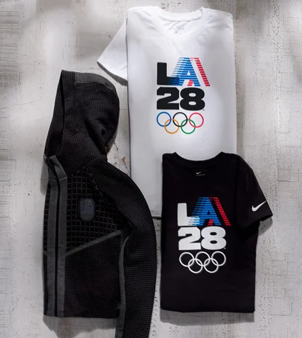 Disposición plana de una sudadera negra LA28, una camiseta blanca con el emblema retro ‘A’ de LA28 y una camiseta negra con el emblema retro ‘A’ de LA28.