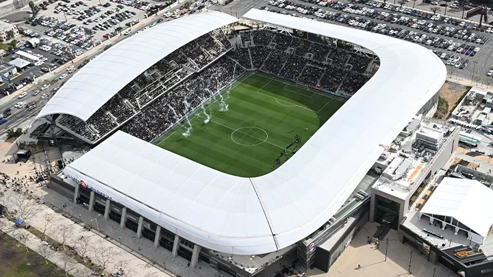 Fotografía aérea que muestra el Stadium in Exposition Park desde arriba, enmarcado por dos aparcamientos alrededor del perímetro. Tres columnas de humo blanco se elevan desde el campo de fútbol con una multitud de espectadores al fondo.