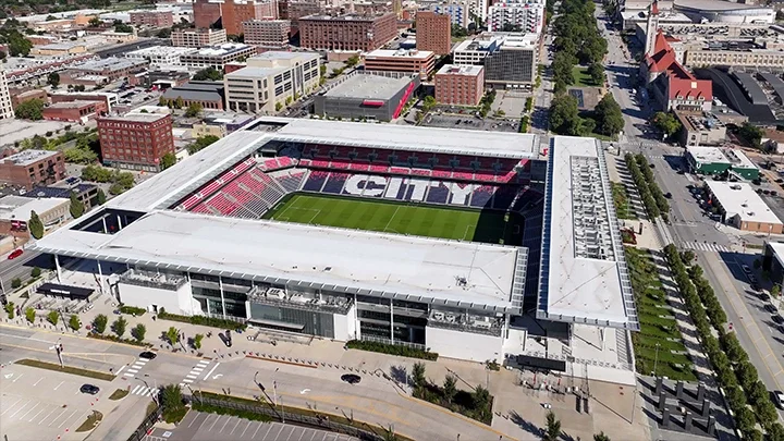 Vista aérea de St. Louis Stadium, con los asientos vacíos del estadio mostrando la palabra “City” en las gradas. Edificios del centro de St. Louis rodean el estadio y, en primer plano, se observa una calle frente a la entrada del estadio.