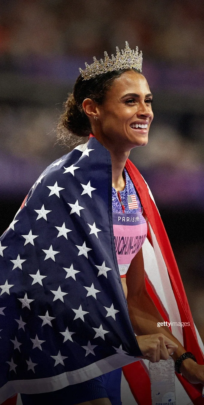 Fotografía de Sydney McLaughlin-Levrone envuelta en la bandera estadounidense, con una corona en la cabeza, tras ganar una medalla de oro en los Juegos de París 2024