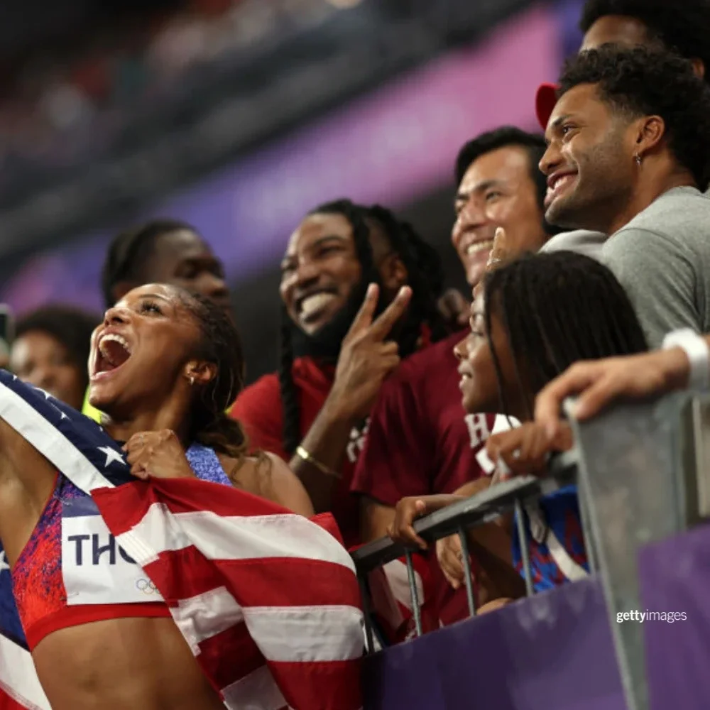 Una fotografía de una atleta femenina de atletismo sonriendo y tomándose una foto con aficionados en las gradas.