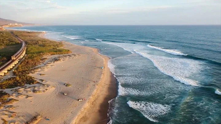 Una fotografía aérea de Trestles Beach en San Clemente muestra el océano a la derecha de la fotografía con arena en la orilla y algunos surfistas llevando sus tablas de surf a la izquierda de la fotografía. En el extremo izquierdo de la fotografía se ve un tren sobre las vías del tren.