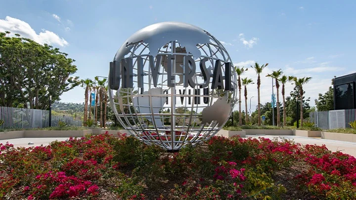 Una fotografía de un globo terráqueo de metal plateado con la palabra Universal en el anverso sobre un lecho de plantas y flores. Al fondo, una fila de palmeras. 