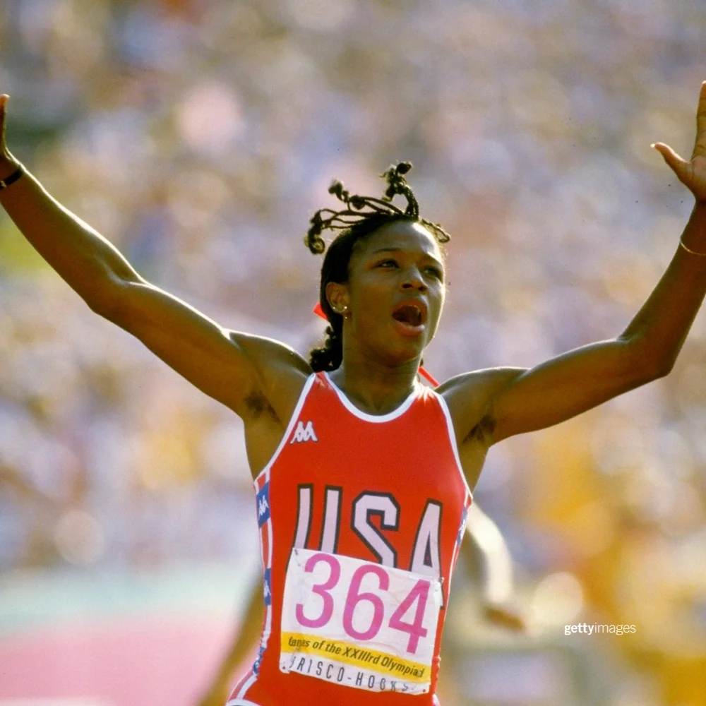 Foto de la atleta olímpica del equipo de atletismo de Estados Unidos, Valerie Brisco-Hooks, celebrando con los brazos en alto, vistiendo un uniforme rojo de atletismo con la palabra USA al frente, mientras corre en la pista al aire libre del Coliseo Memorial de Los Ángeles durante los Juegos de 1984.