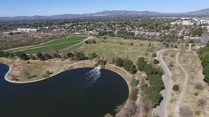 Fotografía aérea de la Zona Recreativa del Río Sepulveda Basin, mostrando un camino pavimentado en primer plano, con césped, árboles y un pequeño lago en la imagen. Al fondo se ven edificios y montañas que representan el Valle de San Fernando.