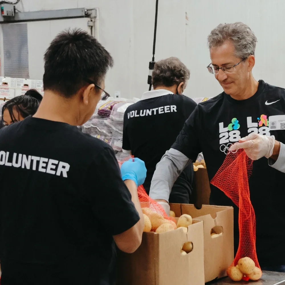Una fotografía de voluntarios clasificando sacos de papas mientras llevan puestos gafetes y camisetas del Programa de Voluntariado LA28